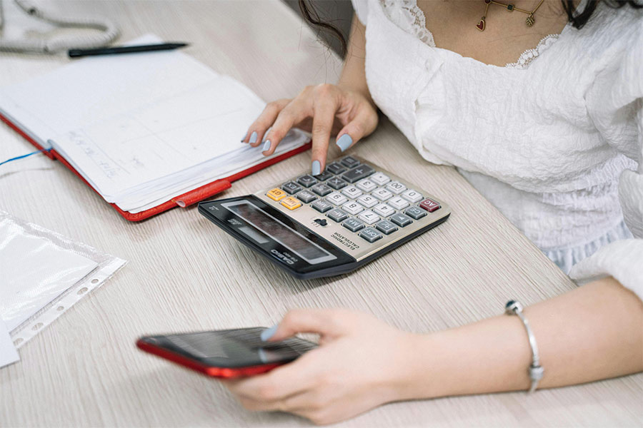 Hire a bookkeeper blog image. Woman using pone, calculator and notebook to manage her accounts. Photo by Anna Tarazevich: https://www.pexels.com/photo/woman-using-a-black-and-red-smartphone-and-calculator-5196820/